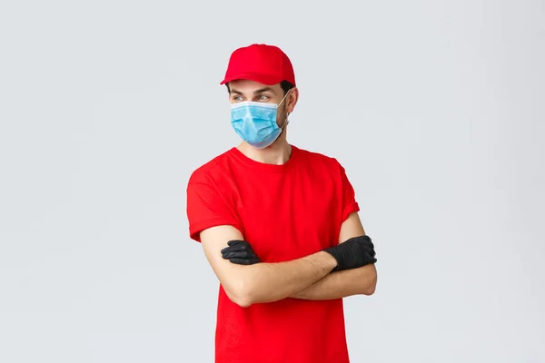 Covid-19, self-quarantena, shopping online e concetto di spedizione. Confidente corriere in uniforme rossa, guanti protettivi e maschera viso, in piedi braccia incrociate, distogliere lo sguardo, pronto prendere i vostri ordini — Foto Stock