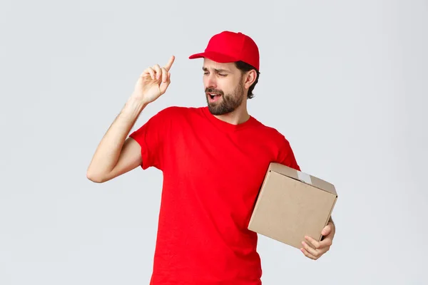 Beställningsleverans, näthandel och paketfrakt koncept. Sorglös vacker skäggig kurir i röd uniform, mössa och t-shirt, innehav box paket, sång sång, grå bakgrund — Stockfoto