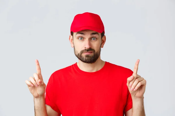 Nätshopping, leverans under karantän- och hämtningskoncept. Obeslutsam och osäker kurir i röd uniform mössa, t-shirt, flin och peka fingrar upp osäker, vet inte, kan inte bestämma — Stockfoto