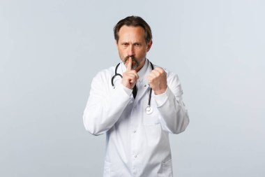 Covid-19, koronavirüs salgını, sağlık çalışanları ve pandemik konsept. Beyaz önlüklü ciddi bir doktor, yumrukla ve susmakla tehdit ediyor, hastanede sessizlik talep ediyor, uyarıda bulunuyor.