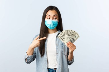 Sosyal mesafeli yaşam tarzı, covid-19 pandemik iş ve heyecan konsepti. Medikal maskeli mutlu Asyalı kız parayı işaret etti ve büyük para buldu.