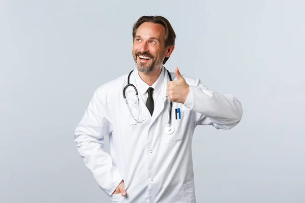 Covid-19, epidemia koronawirusów, pracownicy służby zdrowia i koncepcja pandemii. Zadowolony przystojny uśmiechnięty lekarz w białym płaszczu patrząc w lewy górny róg, pokazać kciuk do góry zatwierdzenia — Zdjęcie stockowe