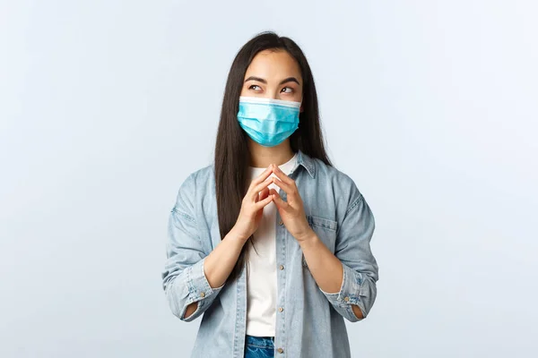 Estilo de vida social distanciante, pandemia covid-19 y concepto de emociones de las personas. Mujer asiática astuta y creativa en máscara médica, plan secreto intrigante, dedos de campanario y mirada soñadora esquina superior izquierda — Foto de Stock