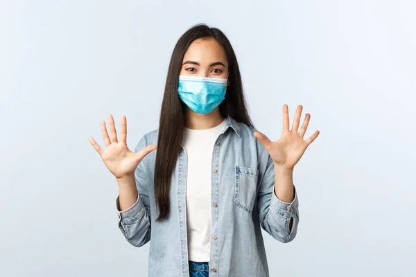 Estilo de vida social distanciamento, covid-19 pandemia e as pessoas emoções conceito. Sorrindo alegre mulher asiática em máscara médica mostrando dez dedos, mãos vazias, fundo branco — Fotografia de Stock