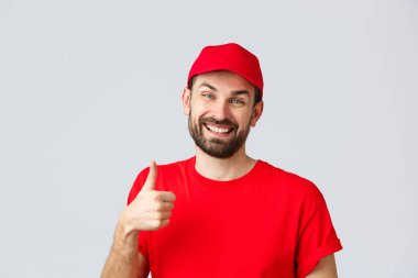 İnternetten alışveriş, karantina sırasında teslimat ve paket servis konsepti. Kırmızı üniformalı, güler yüzlü, sakallı teslimatçı ya da kurye. Destekleyici başparmağını kaldır.