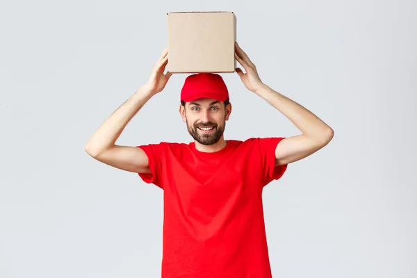 Entrega de pedidos, compras en línea y concepto de envío de paquetes. Divertido y lindo mensajero barbudo en gorra uniforme roja y camiseta, caja de sujeción en la cabeza. Empleado con paquete sonriendo —  Fotos de Stock