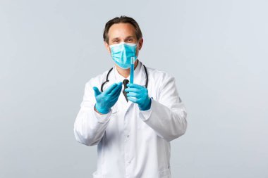 Covid-19, virüsü önleme, sağlık çalışanları ve aşı konsepti. Tıbbi maskeli ve eldivenli tatmin olmuş gülümseyen doktor test tüpünü aşı numunesiyle işaret ediyor, iyi sonuçtan memnun.