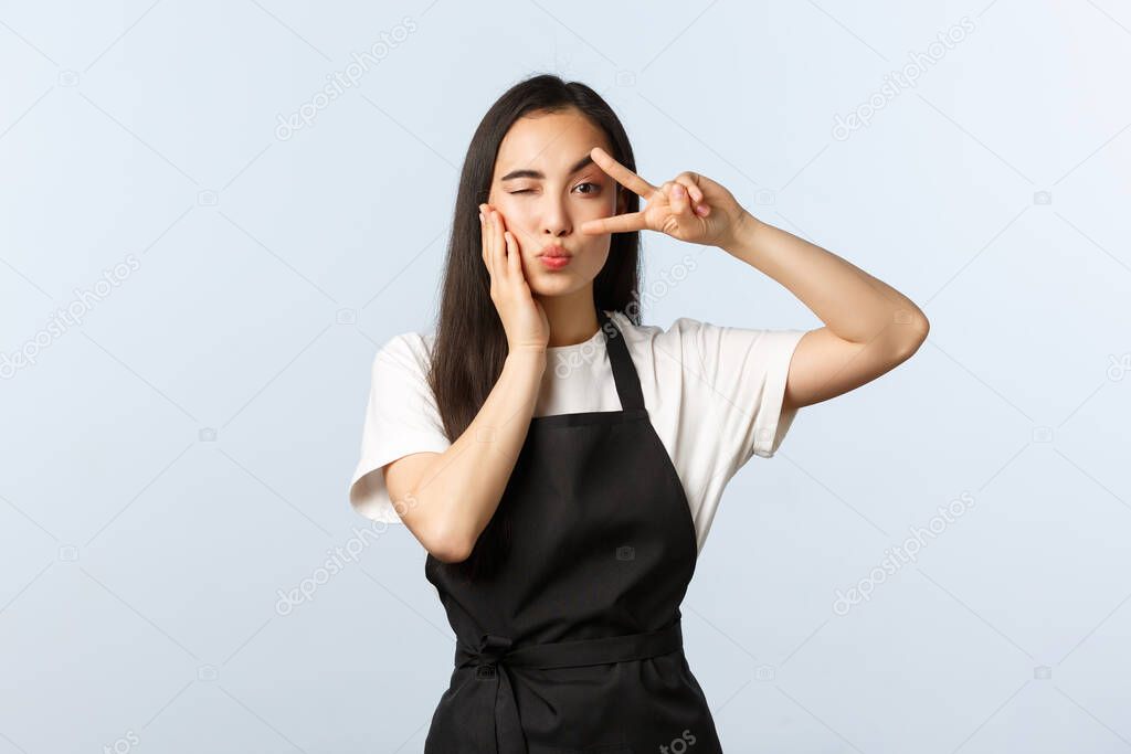 Coffee shop, small business and startup concept. Cute flirty asian female barista inviting to cafe. Attractive cafe staff in black apron, show silly peace sign, wink and fold lips as blowing kiss