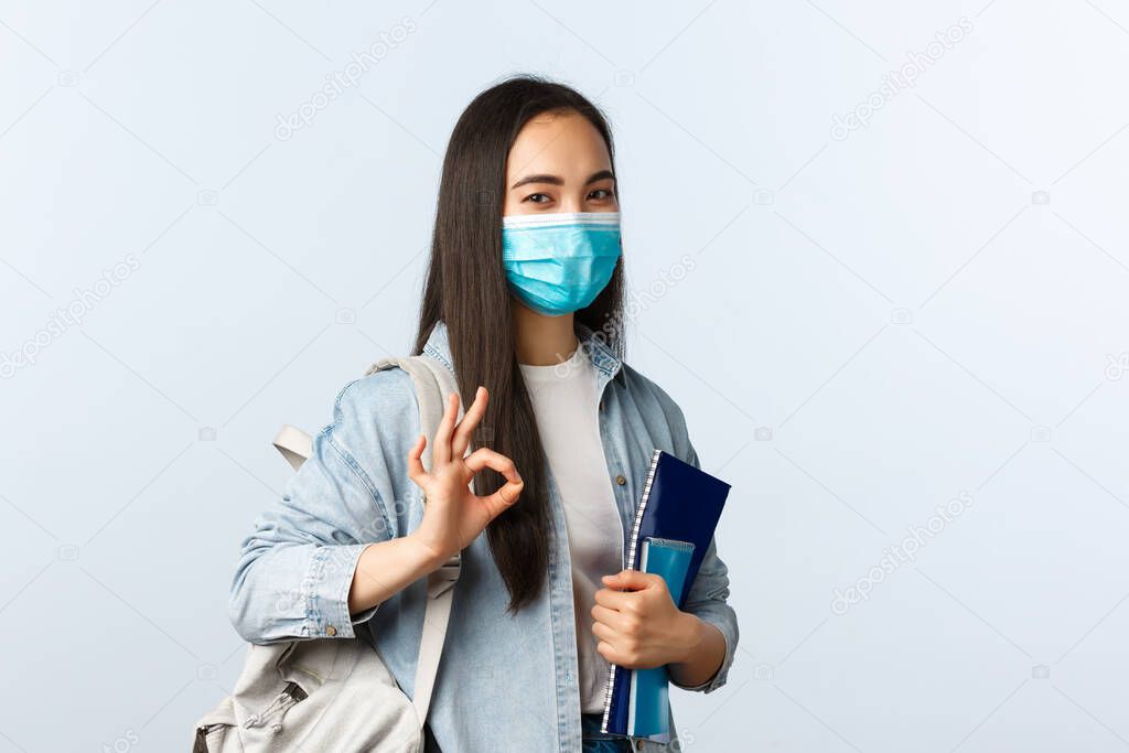 Covid-19 pandemic, education during coronavirus, back to school concept. Satisfied cute asian girl student in medical mask, show okay gesture as studying at cool university, hold notebooks