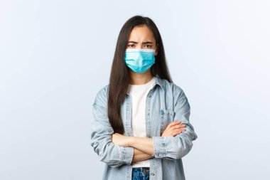 Sosyal mesafeli yaşam tarzı, covid-19 salgın hayatı ve boş zaman kavramı. Şüpheli ve hayal kırıklığına uğramış tıbbi maskeli genç bir kız, Asyalı bir kadın hoşnutsuz görünüyor, çapraz kollar göğsü ve kaşları çatık