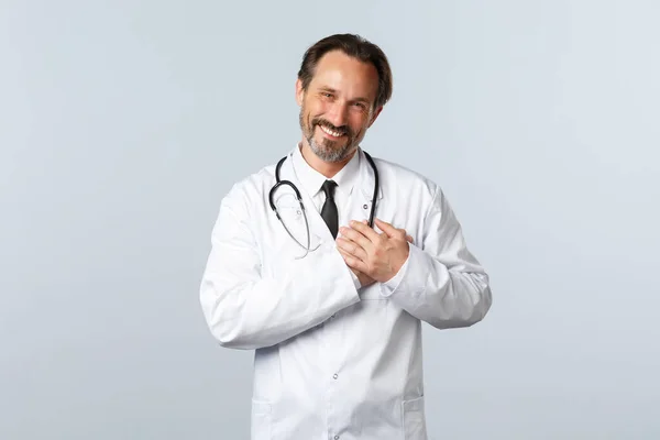 Covid-19, ξέσπασμα του ιού της στέψης, επαγγελματίες υγείας και πανδημία. Συγκινημένος γιατρός να είναι εγκάρδια με ωραία χειρονομία, κρατώντας τα χέρια στην καρδιά και χαμογελώντας ευχαριστημένος — Φωτογραφία Αρχείου