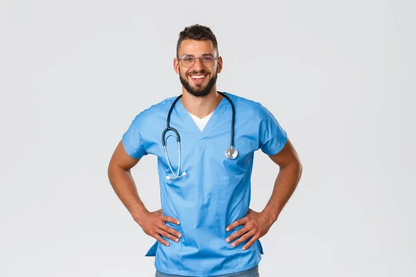 Egészségügyi dolgozók, orvostudomány, kovid-19 és világjárvány önkarantén koncepció. Lelkes jóképű orvos, férfi nővér szemüvegben és kék köpenyben, mosolyog, élvezi a klinikai betegekkel való munkát. — Stock Fotó