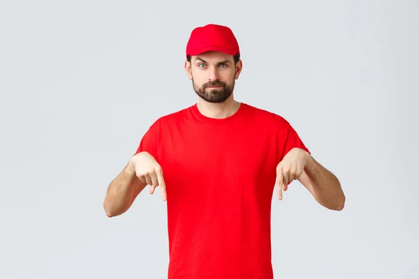 Nätshopping, leverans under karantän- och hämtningskoncept. Skeptisk eller misstänksam kurir i röd uniform mössa och t-shirt, pekar finger ner, stående grå bakgrund — Stockfoto