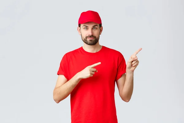 Online winkelen, levering tijdens quarantaine en afhaalconcept. Zelfverzekerde bebaarde koerier in rood uniform pet en t-shirt, glimlachende camera, richting geven, met de vinger naar rechts wijzen, klanten informeren — Stockfoto