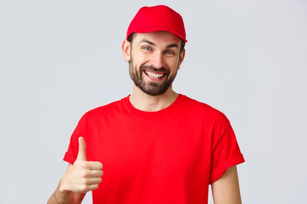 Nätshopping, leverans under karantän- och hämtningskoncept. Vänlig, glad kurir i röd mössa och t-shirt uniform, uppmuntra göra internetbeställningar, tummen upp i godkännande eller rekommendation — Stockfoto