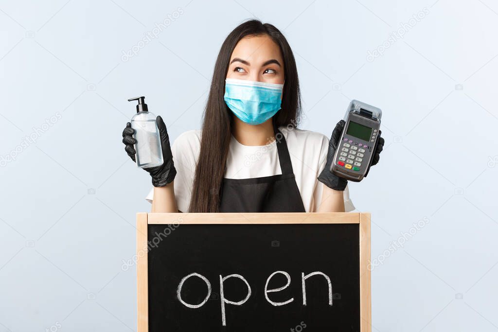 Small business, covid-19 pandemic, preventing virus and contactless orders concept. Excited cute asian waitress, barista in medical mask giggle near open sign, hold hand sanitizer and POS terminal