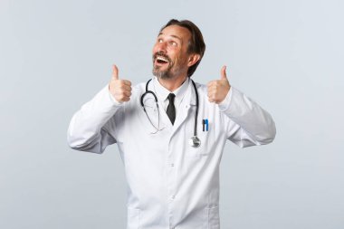 Covid-19, koronavirüs salgını, sağlık çalışanları ve pandemik konsept. Beyaz önlüklü, hevesli, yakışıklı bir doktor sol üstteki reklama bakıyor ve başparmak kaldırıyor.