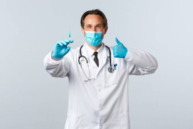 Covid-19, virüsü önleme, sağlık çalışanları ve aşı konsepti. Tıbbi maskeli ve eldivenli güvenilir profesyonel doktor. Baş parmak, Coronavirus aşısı öneriyor, elinde şırınga var.