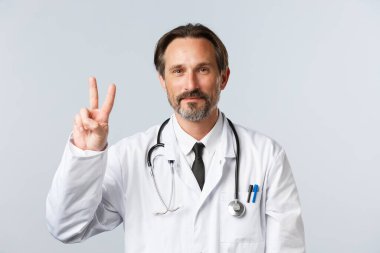 Covid-19, virüsü önleme, sağlık çalışanları ve aşı konsepti. İyimser gülümseyen beyaz önlüklü doktor V-işareti huzuru gösteriyor, hevesli olmak, hasta hastaları tedavi etmek, iyi sonuçlara inanmak