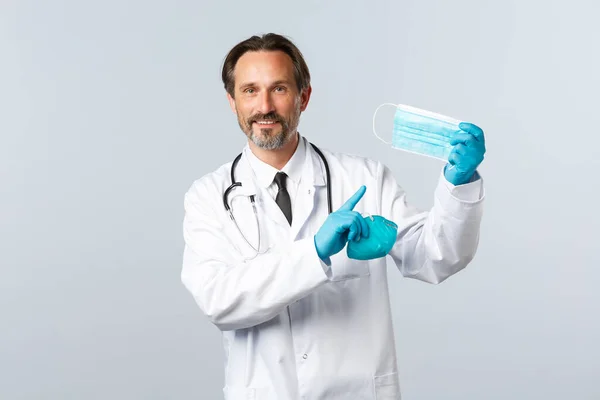 Covid-19, preventing virus, healthcare workers and vaccination concept. Smiling male doctor in white coat and gloves, preventive measures social distancing, hold respirator and point medical mask — Stock Photo, Image