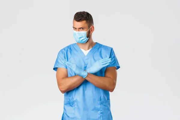 Pekerja kesehatan, covid-19, coronavirus dan konsep virus. Dokter profesional serius, perawat laki-laki dalam pakaian biru dan topeng medis memberitahu berhenti, menunjukkan tanda larangan silang, mengerutkan kening tidak senang — Stok Foto