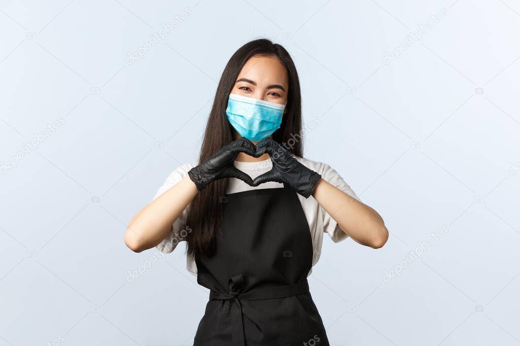 Covid-19, social distancing, small coffee shop business and preventing virus concept. Cute and tender barista, asian female cafe staff care for visitors, show heart sign, wear medical mask and gloves