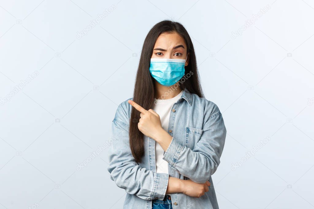 Social distancing lifestyle, covid-19 pandemic everyday life concept. Skeptical displeased asian pretty girl asking explanation, pointing finger left, raise one eyebrow suspicious, wear mask