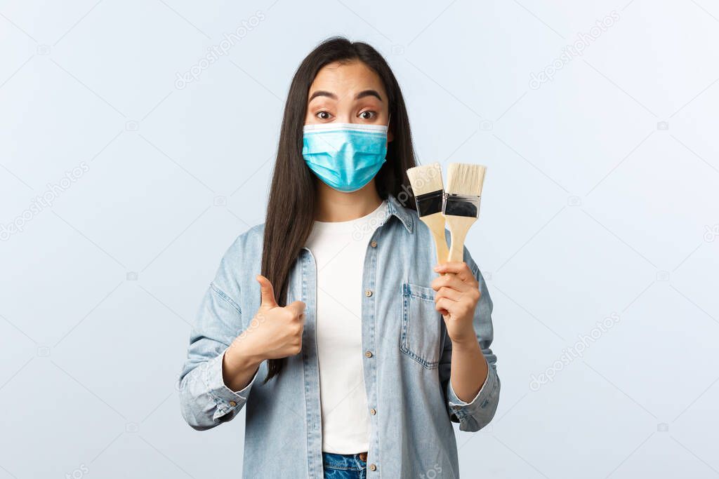 Social distancing lifestyle, covid-19 pandemic, self-isolation hobbies concept. Asian cute woman in medical mask looking satisfied, showing thumbs-up and painting brushes