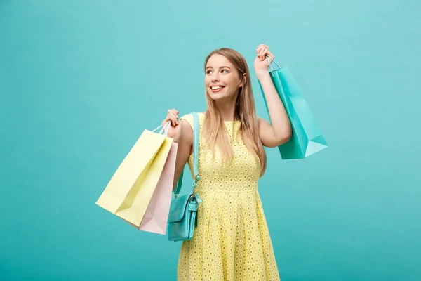 Lifestyle-Konzept: Porträt einer schockierten jungen attraktiven Frau in gelben Sommerkleidern, die mit Einkaufstaschen posiert und vor blauem Hintergrund in die Kamera blickt. — Stockfoto