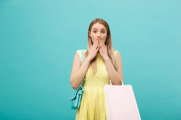 Concepto de estilo de vida: Retrato de una joven atractiva sorprendida en un vestido amarillo de verano con bolsas de compras y mirando a la cámara sobre un fondo azul . — Foto de Stock