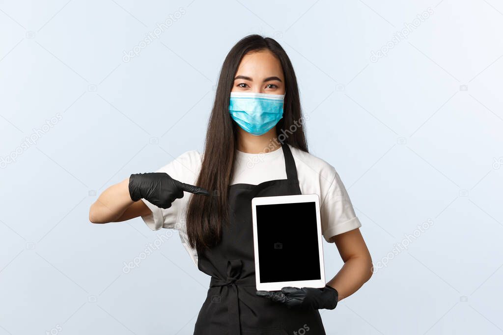 Covid-19, social distancing, small coffee shop business and preventing virus concept. Cute young asian barista, female cafe worker in medical mask pointing at digital tablet