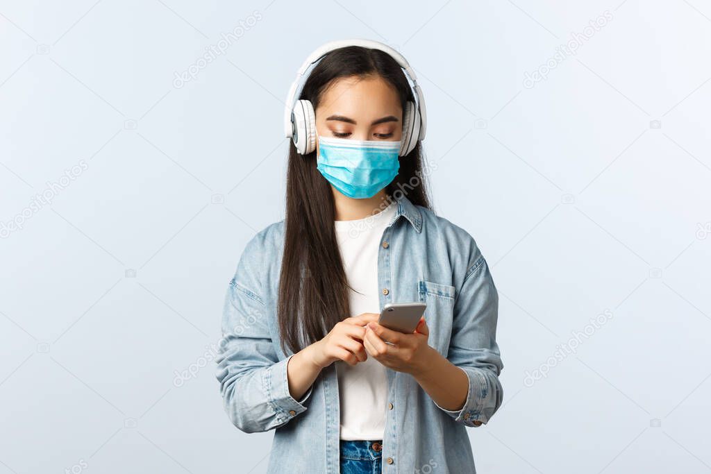 Social distancing lifestyle, covid-19 pandemic and self-isolation leisure concept. Modern cute asian girl in medical mask, listening music wireless headphones, using mobile phone app