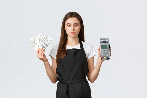 Grocery store employees, small business and coffee shops concept. Saleswoman or barista in black apron holding cash and payment terminal POS, showing two ways of purchasing items in shop