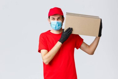 Paketler ve paket teslimatı, karantina teslimatı, nakil emirleri. Dost canlısı kurye müşteri evine sipariş getirdi, paket kutusunu omzuna aldı, maske taktı ve lastik eldiven taktı.