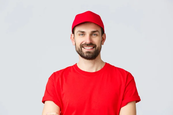 Achats en ligne, livraison en quarantaine et concept à emporter. Beau courrier barbu en uniforme rouge, souriant gai, poitrine croisée, prêt à livrer votre commande, fond gris — Photo