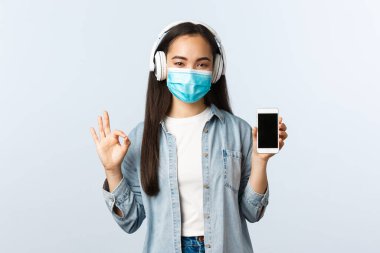 Sosyal mesafeli yaşam tarzı, covid-19 salgını ve kendini izole etme eğlence konsepti. Tatmin olmuş gülümseyen tıbbi maskeli tatlı Asyalı kız, müzik kulaklıkları dinle, tamam işareti ve akıllı telefon ekranını göster.