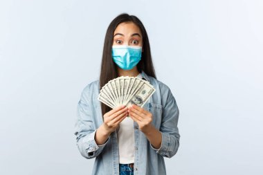 Sosyal mesafeli yaşam tarzı, covid-19 pandemik iş ve heyecan konsepti. Medikal maskeli mutlu Asyalı kadın elinde para tutarak evinden online iş için para aldı.