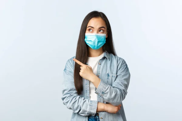 Estilo de vida social distanciamento, covid-19 pandemia vida cotidiana e conceito de lazer. Intrigado muito asiático mulher no médico máscara anunciar banner, apontando dedo esquerdo — Fotografia de Stock