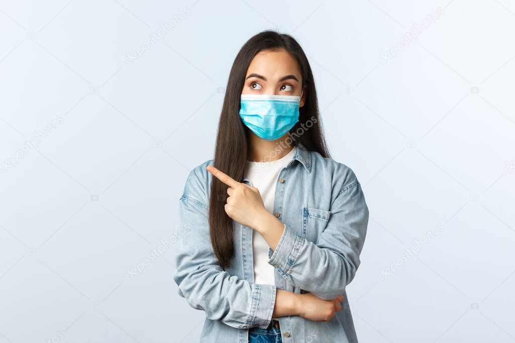 Social distancing lifestyle, covid-19 pandemic everyday life and leisure concept. Intrigued pretty asian woman in medical mask advertise banner, pointing finger left