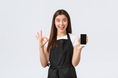 Market çalışanları, küçük işletmeler ve kahve dükkanları konsepti. Heyecanlı siyah önlüklü bayan barista akıllı telefon görüntüleme uygulaması gösteren iyi bir kullanıcı deneyimi garanti ediyor, tamam işareti yap
