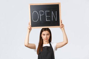 Market çalışanları, küçük işletmeler ve kahve dükkanları konsepti. Siyah önlüklü, ciddi görünümlü genç bir barista açık tabela kaldırın, müşterileri covid salgınından sonra çalışan dükkan veya kafe hakkında bilgilendirin.