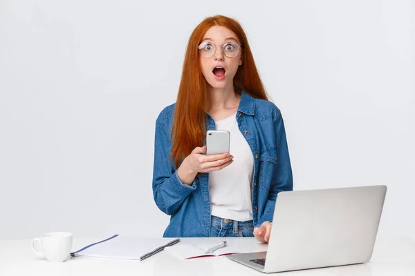 Sorprendido, sorprendido y sacudido adolescente pelirroja mirando sin palabras cámara, caída de la mandíbula jadeando mirada maravillada, leer noticias fascinantes, consiguió mensaje en el teléfono inteligente, de pie cerca de la computadora portátil y escritorio — Foto de Stock