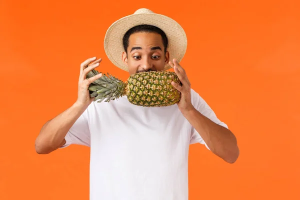 Funny handsome hipster guy, african american tourist enjoying vacation, trying bite pineapple, looking at fruit, standing orange background joyful, like travel hot countries during winter season — 스톡 사진