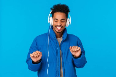 Guy catching positive vibes, feeling groovie and satisfied with new headphones. Cheerful african american guy wearing winter padded jacket, bought earphones holidays sale, dancing and listen music
