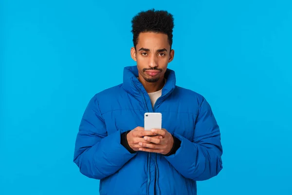 Расстроенный и разочарованный обеспокоенный африканский американец молодой парень упустил шанс, чувствуя себя несчастным и угрюмым, хмурясь, держа смартфон, стоя обитой зимней курткой синий фон — стоковое фото