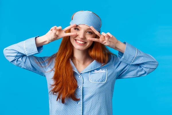 Energized cheerful girl slept well, wake up morning enthusiastic and excited, showing peace signs feeling optimistic smiling toothy, wearing sleep mask and nightwear, standing blue background