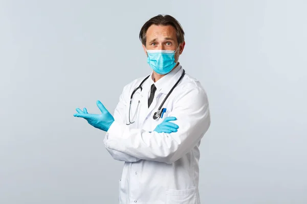 Covid-19, preventing virus, healthcare workers and vaccination concept. Skeptical confused doctor in medical mask and gloves pointing disappointed left, express frustration and disagreement