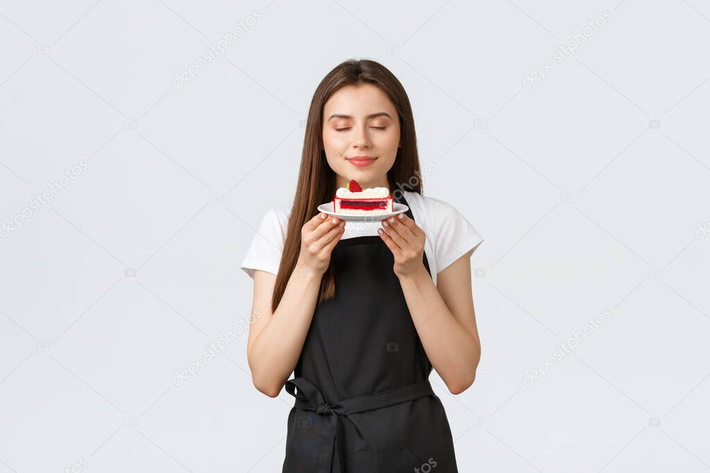 Grocery store employees, small business and coffee shops concept. Female cafe worker in black apron holding tasty cake and smelling delicious dessert with pleased smile and closed eyes