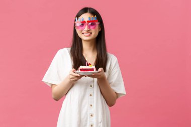 Kutlama, parti tatili ve eğlence konsepti. Gülen kawaii Koreli komik güneş gözlüklü kız elinde doğum günü pastası, kutlama keyfi, pembe arka plan neşeli