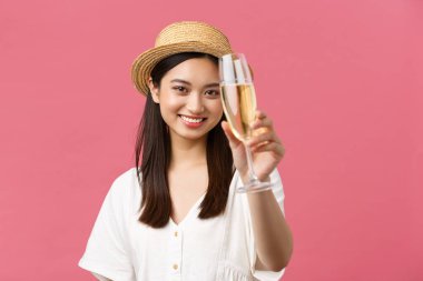 Güzellik, duygular, rahatlık ve tatil konsepti. Hasır şapkalı, neşeli, gevşemiş kadın turistin yakın çekimi, mükemmel yolculuk için cam şampanya kaldırma, şerefe, pembe arka planda dikilme